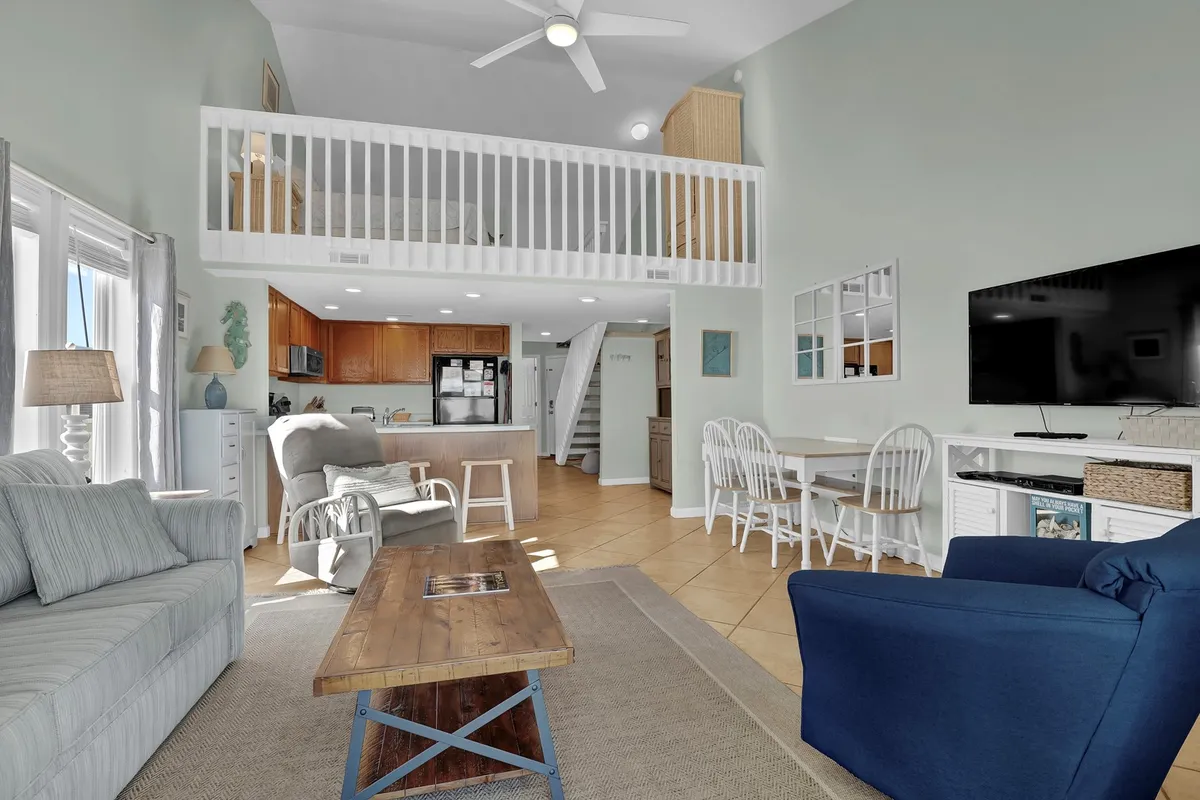 A welcoming living room scene with a comfy sofa, dining area, kitchen, and staircase in view. The space is designed for relaxing, dining, and enjoying time together, bathed in warm, natural light. The arrangement invites gatherings for games, meals, or lounging.
