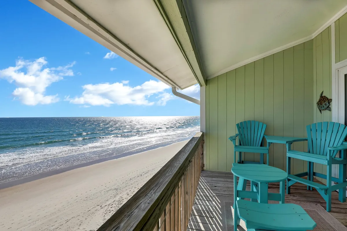 An exterior shot of a beachfront scene with turquoise chairs on a deck overlooking the sandy beach and shimmering ocean. The sky is clear and blue, enhancing the sense of openness and calm. The setup evokes a relaxed atmosphere, ideal for unwinding and savoring the natural beauty.