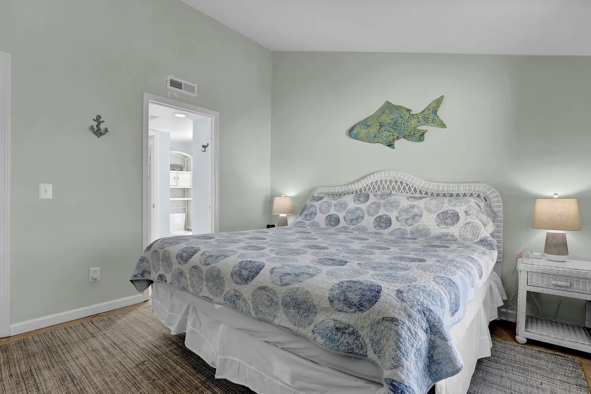 A tranquil bedroom scene featuring a bed with a stylish patterned quilt, fish-themed wall art, and nightstands with lamps on either side. Sunlight filters softly into the room, enhancing its restful atmosphere.