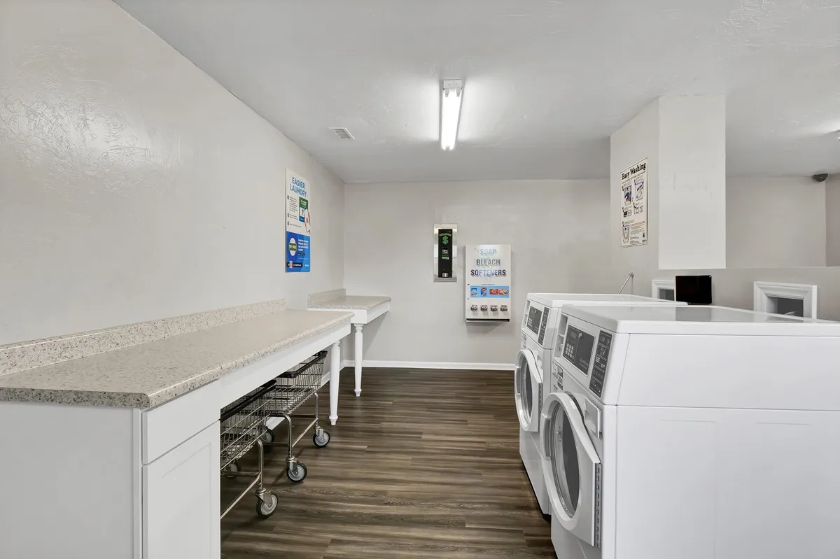 A practical, organized laundry room interior, featuring several washing machines, a countertop for folding clothes, and laundry carts. The layout is clean and efficient, emphasizing the convenience of on-site laundry facilities.