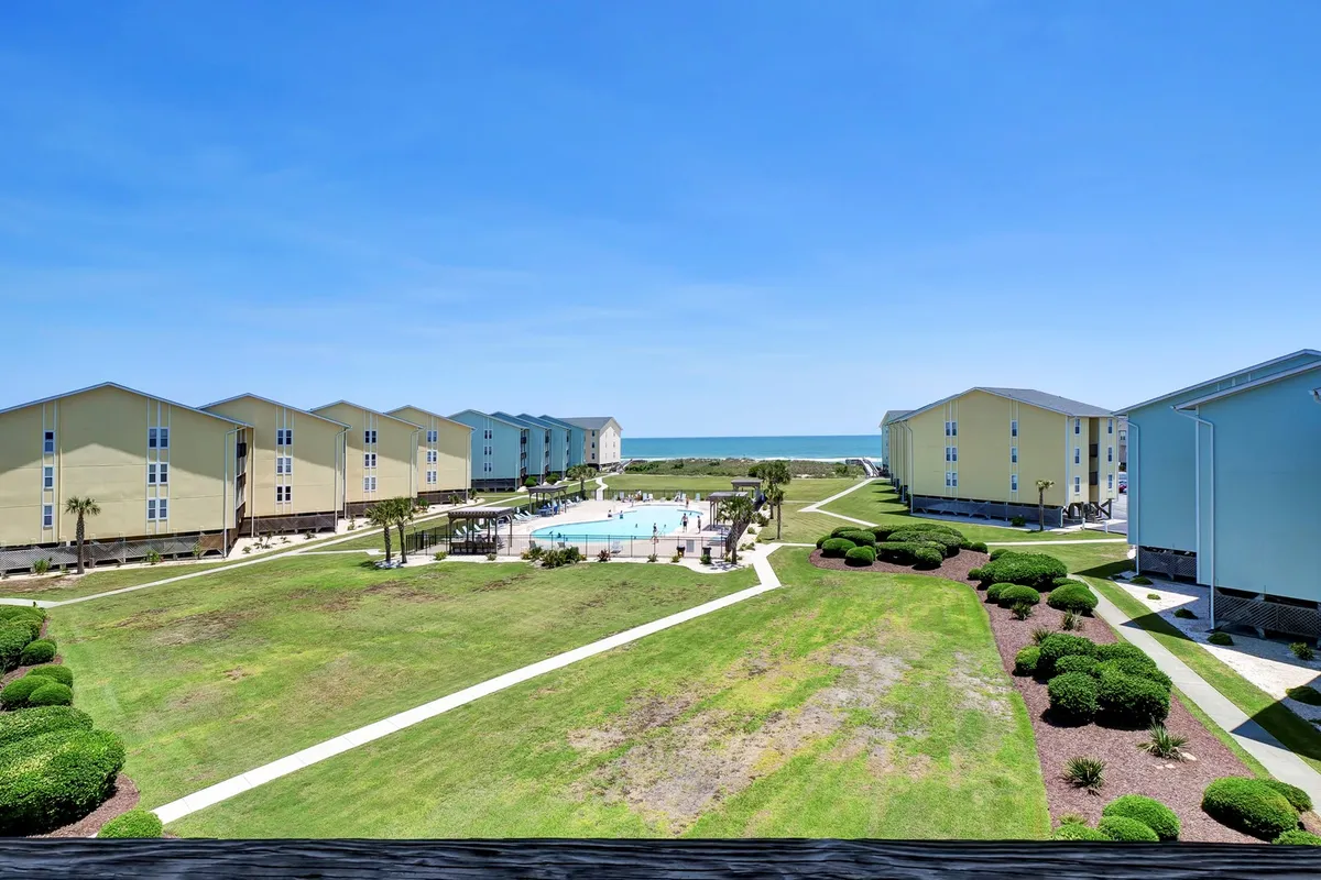 This exterior image captures the community's swimming pool framed by a green lawn, with views of the beach and nearby buildings. The pool area is bathed in natural light, inviting guests to relax, swim, or socialize in a laid-back coastal setting.
