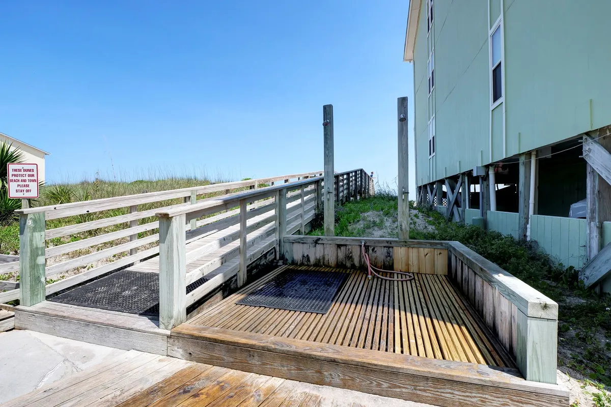 A wooden deck area with a refreshing beach shower, surrounded by grass and a path leading directly to the beach. The deck is bathed in natural light, suggesting a casual, relaxed environment perfect for rinsing off after seaside adventures together.