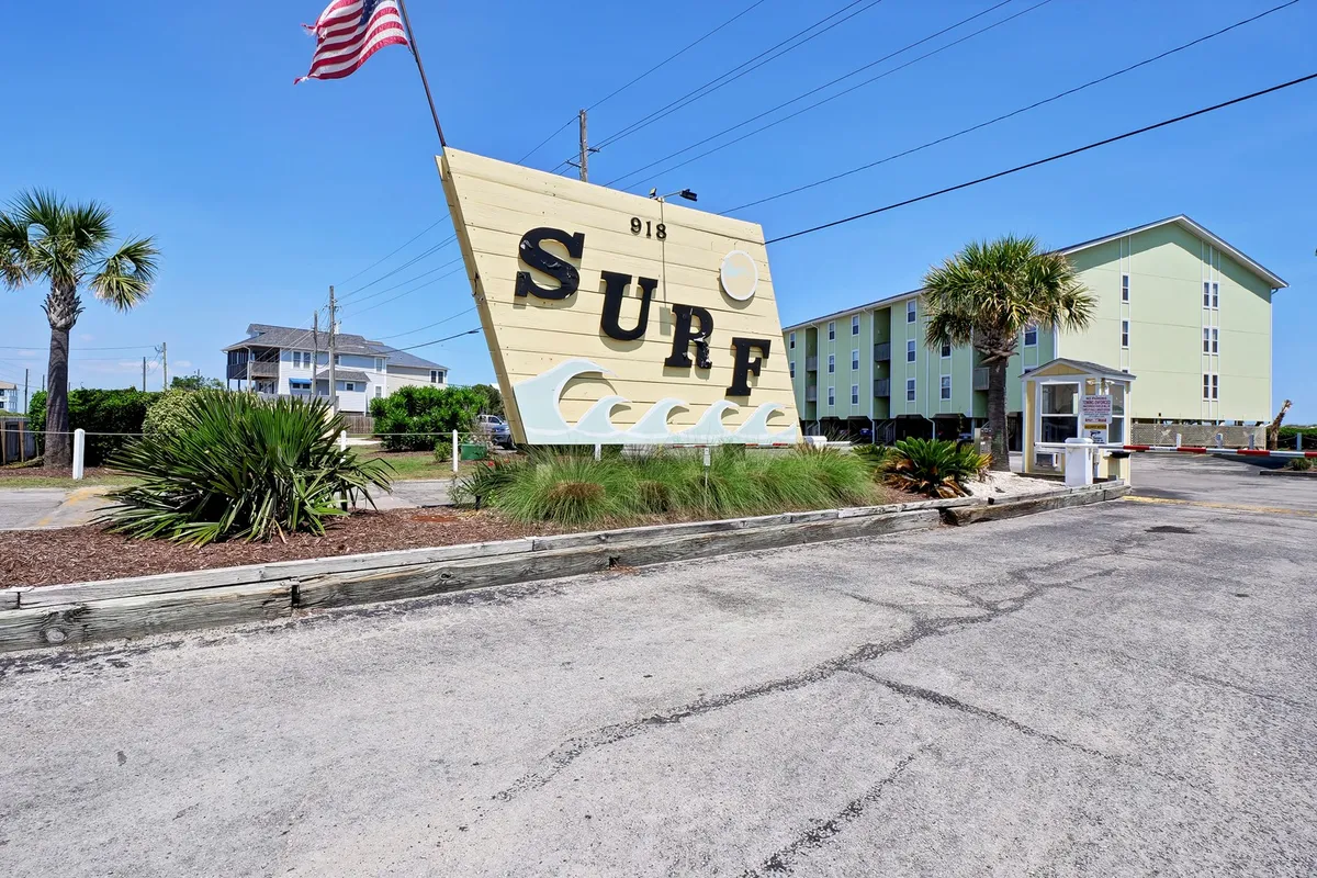 A vibrant exterior shot featuring a large surf sign, palm trees, and surrounding buildings. The scene is bright and lively, evoking the energy and excitement of Surf City just steps away from the condo.