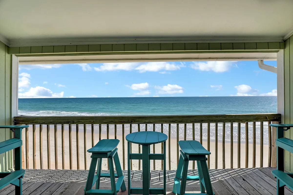 The image showcases a tranquil balcony scene overlooking the beach and ocean, with comfortable chairs and a table inviting guests to sit and take in panoramic water views. The soft light and open sky suggest an ideal setting for watching sunrises or enjoying evening cocktails while listening to the waves.