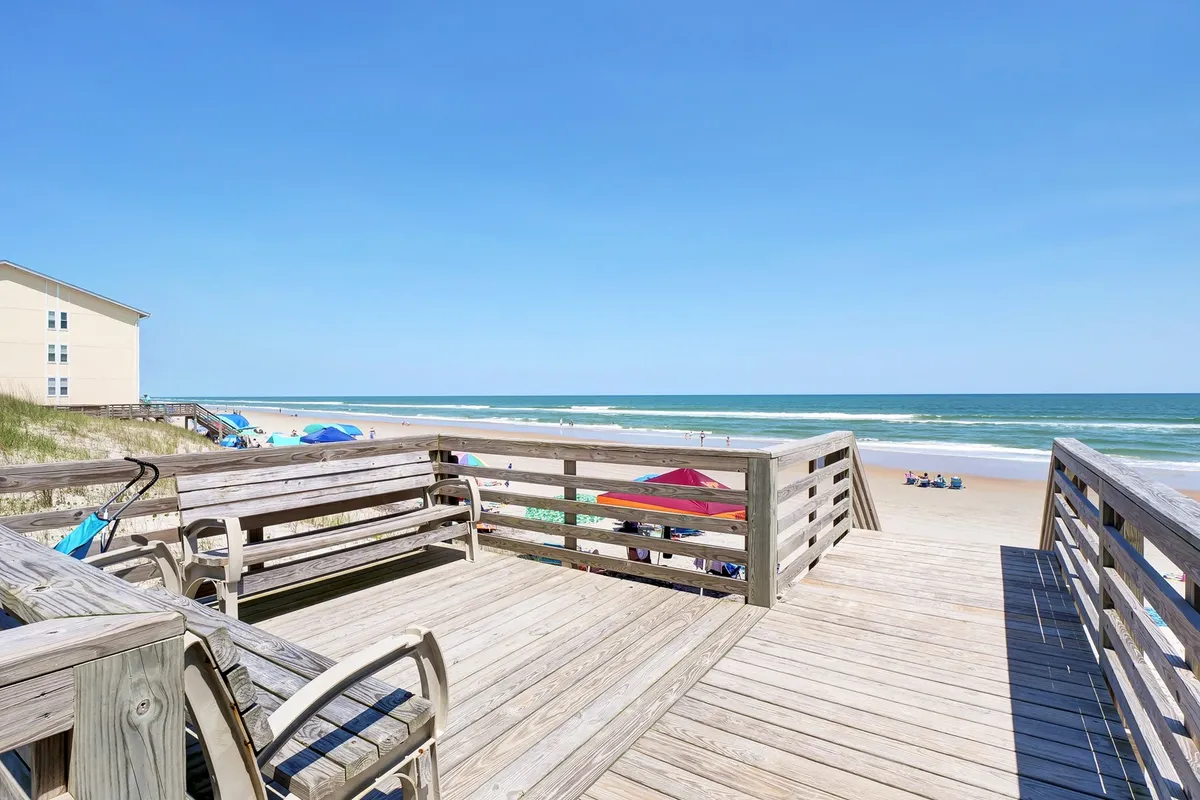 An inviting beachfront exterior with a wooden deck, beach chairs, and umbrellas, opening out to sandy shores and the ocean beyond. The scene suggests cheerful leisure, group activities, and the opportunity to enjoy both sun and stars.