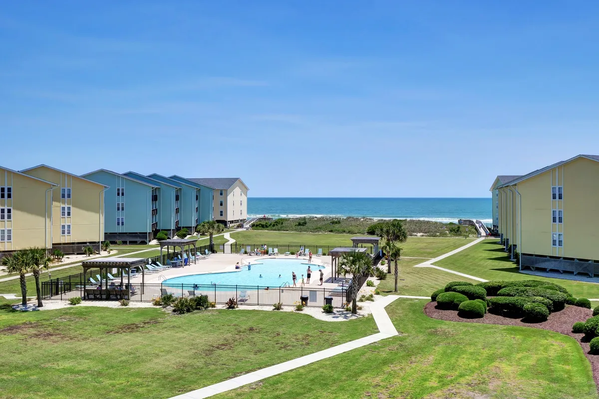 A shared swimming pool overlooks the beach, with the ocean and beachfront buildings visible in the background. The inviting pool area suggests a place for families to swim, sunbathe, and unwind after a day on the sand.