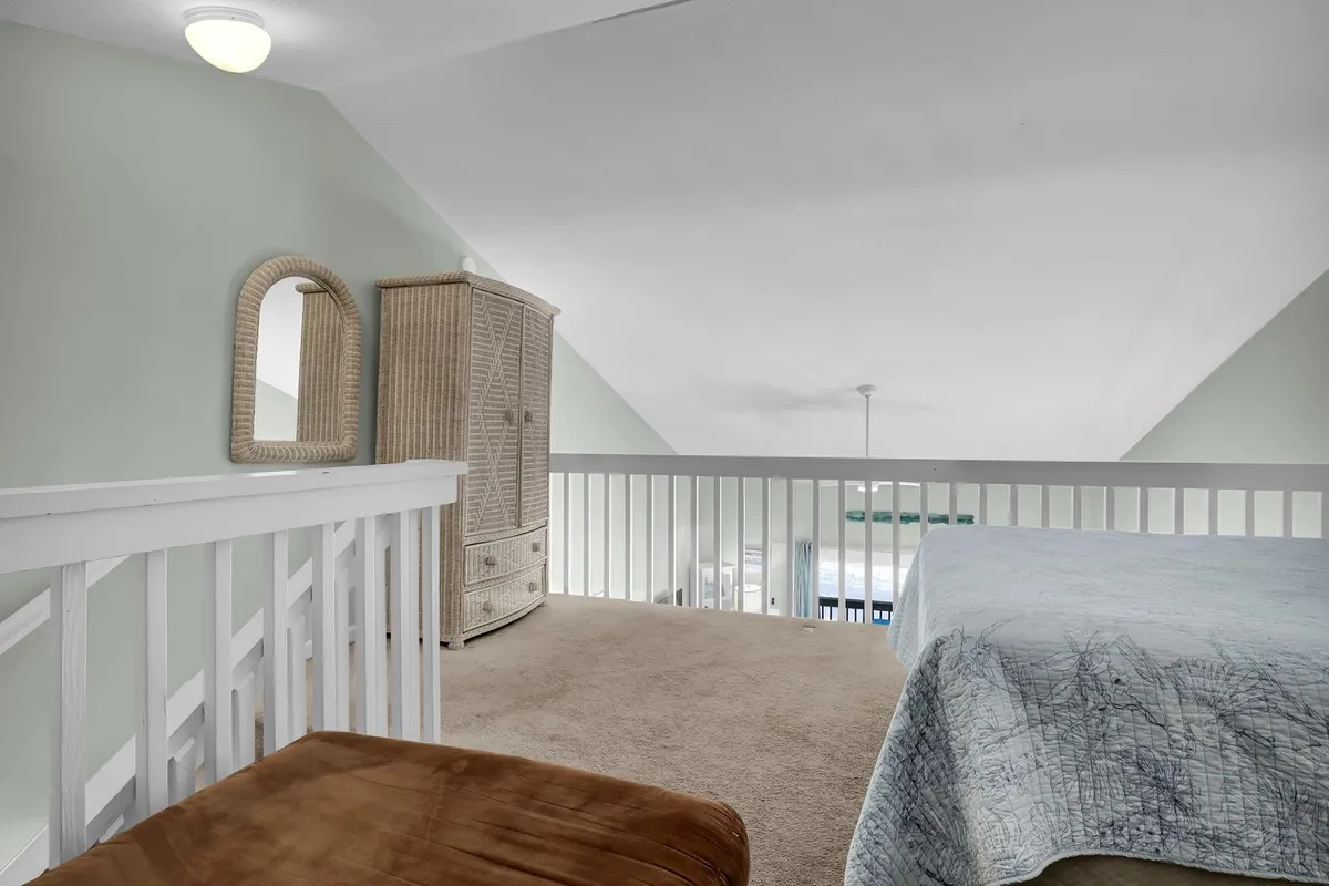 A bright loft space with a lofted ceiling, a cozy bed, a rattan cabinet, and a wall mirror. The open layout and natural light create an inviting, airy ambiance ideal for relaxation.
