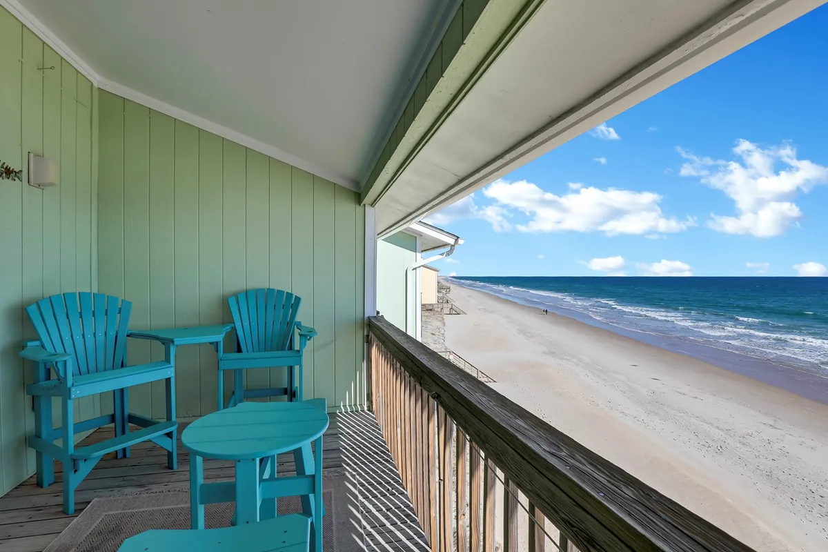 A private balcony featuring blue Adirondack chairs arranged to face a sweeping ocean view, bordered by a wooden railing. The perspective suggests an elevated vantage point, perfect for taking in the tranquil scenery of the beach and horizon. Sunlight pours in, highlighting the inviting seating area and the open sky beyond.